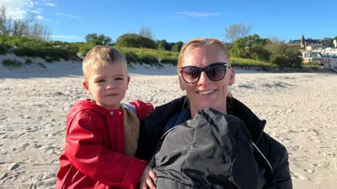 Catherine Richardson wears dark sunglasses and holds her young son Max dressed in a red jacket on a sandy beach. Behind them are grassy sand dunes, scattered trees, and a row of light-coloured buildings, with a bright blue sky and thin, wispy clouds overhead.