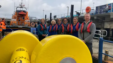 Two large yellow buoys laying on the ground. There are six people standing behind them smiling - one man and five women. In the background there is a orange RNL lifeboat