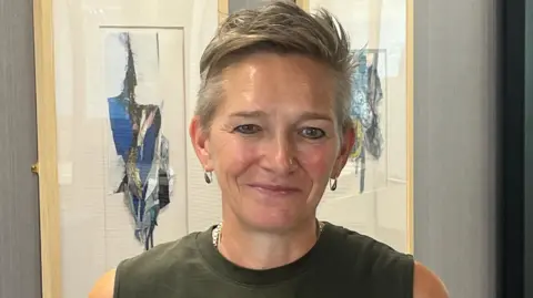 A woman with short light hair and wearing a khaki top in front of art work.