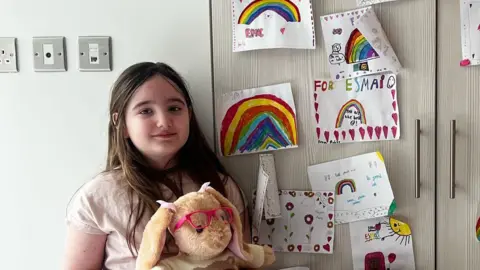 PA Real Life Esmai is stood in front of a set of doors which have pieces of paper attached to them with hand-drawn rainbows. She is holding a rabbit cuddly toy which is wearing pink-framed glasses.