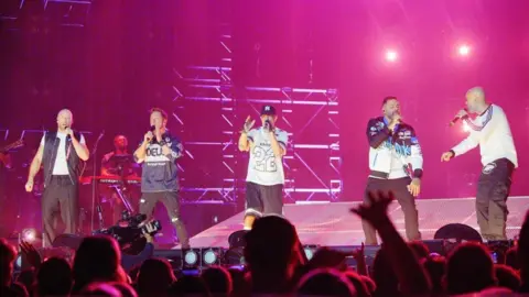 Five members of the boy band are on a bright pink lighted stage. Each of the members are holding a microphone and appear to be singing. There are people on the bottom of the image watching and a man with a camera filming. 