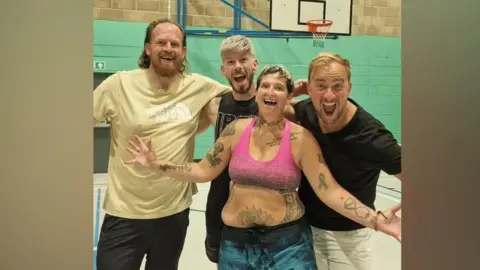 Contributed Four people standing together and smiling at the camera with their arms out. One man is wearing a cream t-shirt with long hair and a beard. Next to him is a blond haired person wearing a black tank top. Standing in the foreground is a woman wearing a pink sports bra with tattoos over her arms and torso. Next to a man wearing a black t shirt and white trousers. 