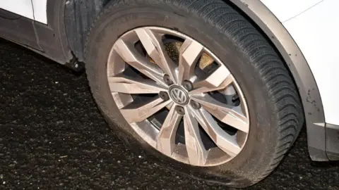A close up shot shows a tyre of a white Volkswagen clearly flat. 