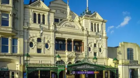 Manx Scenes An ornate, Victorian-era theatre with a cream frontage. "Gaiety Theatre and opera house" is written in letters on the third floor, along with a greek statue of a woman holding a touch aloft. 
On the ground level there is a steel and glass overhanging shelter above the entrance. 