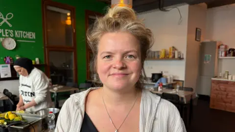 A woman with blonde hair tied in a messy bun stands smiling at the camera in a community kitchen or café, wearing a black top, light cardigan, and silver necklace. Another person prepares food in the background.