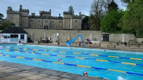 Swimmers lane swim in an outdoor pool. There is a blue water slide and picnic benches on the patio. On the other side of the wall is a castle-like country house with trees surrounding it. The sky is grey and cloudy.