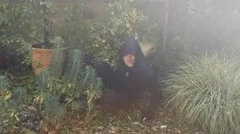 Kent Police A man wearing a black hooded jacket lying on the ground among some plants and small hedges. He is looking into the camera.