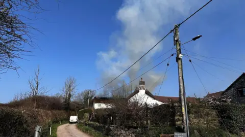 Smoke rises from a house with a lane leading to it and an overhead cable.