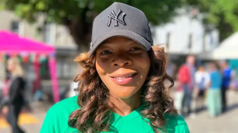 Close-up portrait of a Lainah who is wearing a black baseball cap with a embroidered “NY” logo and a green T-shirt, standing outdoors in a sunlit public space with trees, tents, and blurred people in the background