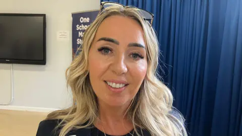 A blonde woman with blue eyes and sunglasses on top of her head smiles at the camera.