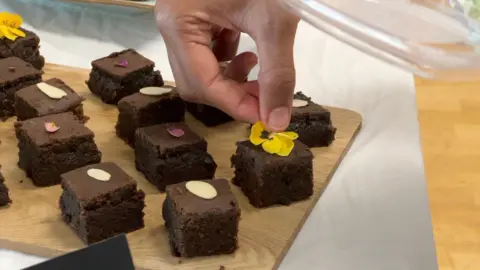 About a dozen chocolate brownie squares, each topped with a petal, on a wooden tray.