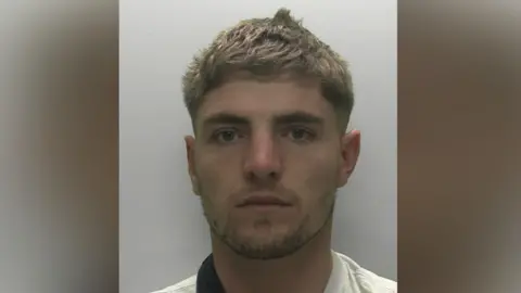 A young blond man with a white top looks at the camera.