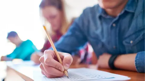Getty Images A student in non-school uniform holds a pencil and writes on a quiz sheet. Their face cannot be seen and other students behind him are blurred.