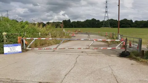 Google A rural access road is blocked by a red-and-white metal barrier gate secured across the entrance. Traffic cones line the road beyond the gate, leading into open fields. A small sign on the left warns against unauthorised access, and hedges and vegetation border the entrance. In the background, a large electricity pylon and overhead power lines stretch across a cloudy sky.
Provide your feedback on BizChat