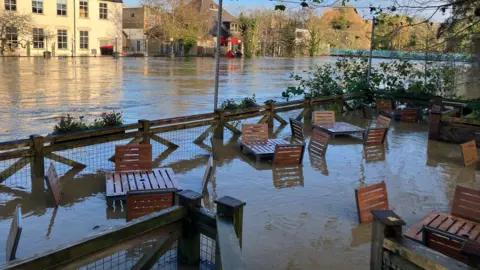 Chairs and tables are completely submerged in murky flood water after the River Avon burst its banks. You can see flood water behind which stretches across the town.