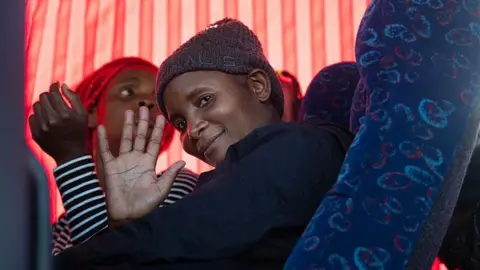 A woman in a grey hat sitting in a coach smiles and waves from the window at the people outside. 