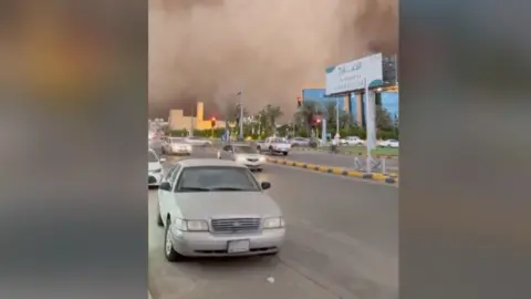 Cars drive down road as huge sandstorm cloud looms in the background 