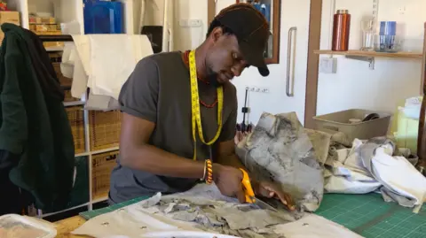 A young, black man, wearing a black cap and a grey T shirt with a red beaded necklace on and a yellow and red striped bracelet. He has a yellow measuring tape wrapped around his neck, and is cutting a piece of grey, tie-dyed fabric with a pair of orange scissors. He is standing at a table with other bits of fabric placed across it.