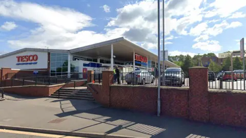 Getty Images A view of Tesco superstore from the road. The store has a white roof and glass front and has Tesco in red capital letters on both the front and side. There is a brick wall around the side with black railings.