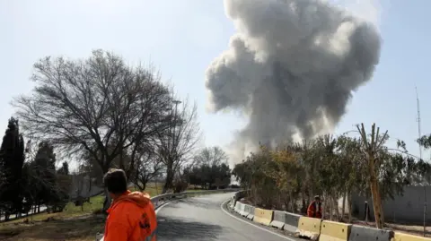 Reuters A workman in high-viz jacket stands in the middle of a road with his back to the camera watching a plume of smoke 