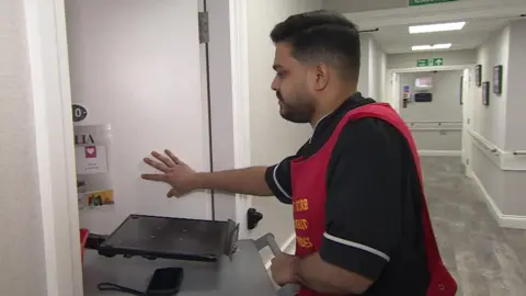 BBC Mohan Sivashankar, a care worker wheeling a trolley into a room in a care home. He is wearing a navy top and a red apron. He has short, black hair and a beard.