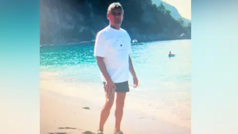 Supplied Nic on a beach on holiday in the past. He is wearing a white t shirt and some shorts. 