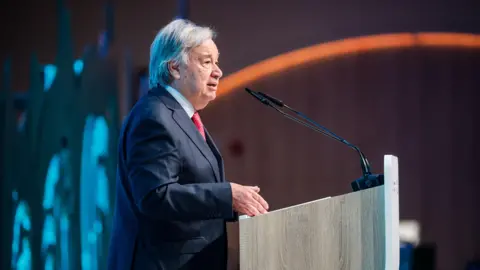 EPA-EFE/REX/Shutterstock United Nations Secretary-General Antonio Guterres stands at a lectern. He is wearing a dark blue suit and a red tie.