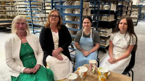 BBC Four women sitting inside a pottery factory with shelves stacked with pottery behind them. It is four generations of the same family, with an older woman with light white hair on the left. On the right is a young woman with long brown hair. In between are two women who are both wearing glasses and have long hair, with one wearing a light blue apron.
