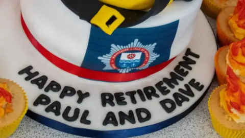 HIWFRS A close up of a cake with inscription 'Happy retirement Paul and Dave'. The cake is in the form of a firefighter helmet, with the Hampshire fire service logo on it. Cupcakes have also been arranged around the cake.