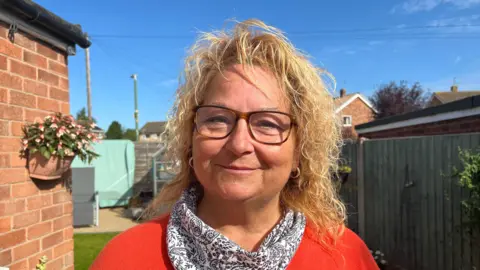 BBC A woman in a white, paisley-patterned scarf and orange jumper standing in a garden