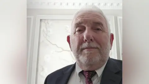 Phil Waring Phil Waring looking into the camera. He is wearing a suit and is stood in front of a white wall.