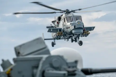 Crown Copyright A grey Royal Navy Wildcat helicopter in flight, equipped with the Martlet missile system. The picture is a close up and the helicopter's rotor's are spinning. 