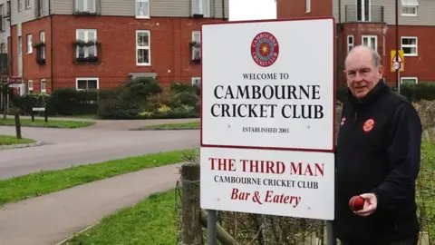 Brian Farmer/BBC A man dressed in a dark top holding a cricket ball. He is standing next to a sign which says: "Welcome to Cambourne Cricket Club, established 2003, The Third Man, Cambourne Cricket Club, Bar & Eatery".