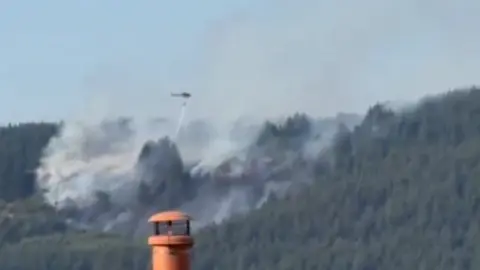 A helicopter in the distance drops water on the woodland and grass fire in Macclesfield Forest, smoke is visable.