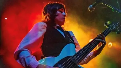 CHERYM Emer McLaughlin has short brown hair and wears a waistcoat, tie and white shirt.  She holds a guitar in her hands while the background is full of coloured smoke. 