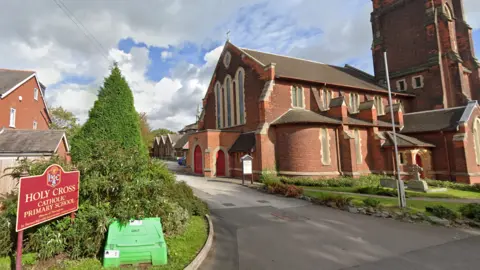 A sign for Holy Cross school to the left. The school building can be seen in the background behind a red-brick church.