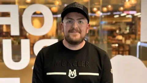 A man wearing a black Moncler sweatshirt, black cap and clear-framed glasses stands with his hands clasped in front of him. He has a short beard. He is standing in front of a glass window with large white letters, with a warmly lit restaurant visible behind him. 