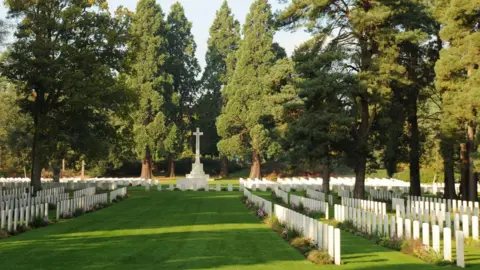 Commonwealth War Graves Commission A shaded green lawn with rows of square white headstones, a large stone cross monument, and tall green trees behind. 