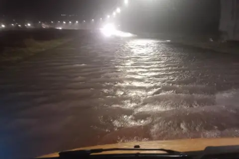 View of a flooded road at night taken from inside a vehicle. The white bonnet and windscreen wipers are visible at the bottom of the screen. Deep water lit up by street lights stretches out into the distance.
