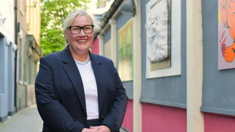 Newcastle City Council leader Karen Kilgour stands next to an art display outside smiling. She is wearing glasses and has blonde hair.