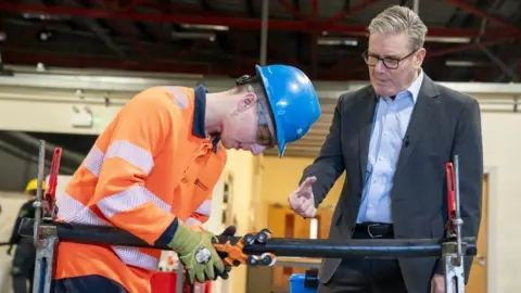 PA Media Keir Starmer, who has grey hair and glasses, speaks to a young industrial worker in an orange high-visibility top and blue hardhat. 