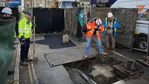 Penzance Council The picture shows a street excavation site. The scene includes a large open trench in the road surface, exposing pipes and a section of granite beneath the tarmac. In the trench is a large equal-sided cross. Two workers in high-visibility jackets and hard hats are closely inspecting and photographing the exposed area, while others stand nearby with tools. Safety barriers and fencing surround the site, and construction equipment and vehicles are visible in the background along with nearby shops. 
