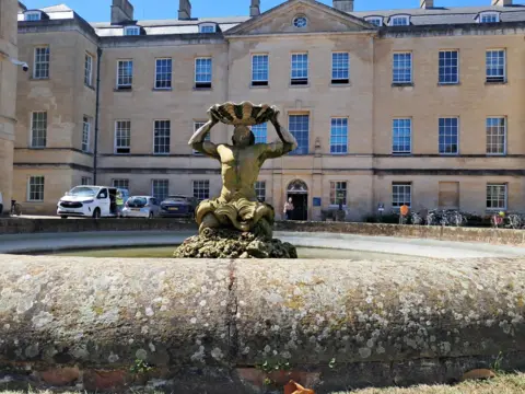 BBC/Molly Pipe A statue of a man holding a large shell above his head stands in a fountain in front of a grand stone building.