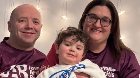 Dean and Becki are smiling wearing red / maroon football style Juvenile Arthritis Research. She has long brown hair and glasses. They are both sat on a grey sofa. She is holding their son who has brown hair, a white top with blue writing and grey and blue striped trousers.