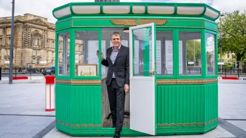 LDRS Image shows Steve Rotheram, in a suit, stepping out of a green Art Deco toll booth. 