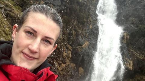 Ruth Trinder standing in front of a waterfall. Her dark hair is pulled back behind her head, she has small hoops in her ears and is giving a slight smile to the camera. She wears a red and black jacket