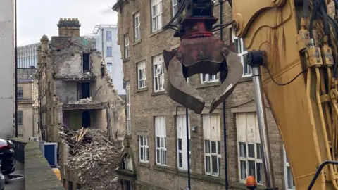 A collapsed building in the background and the yellow metal arm of a mechanical pincer in the foreground 