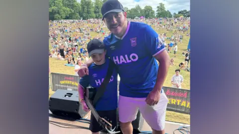 Ollie Ward/BBC A young boy and man pose together on a stage and smile at the camera. They wear matching blue Ipswich Town football shirts. The boy has an acoustic guitar around his torso and he is wearing a black cap and shorts. The man is wearing a blue hat as well as white shorts. Crowds below the stage in a park watch on.