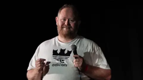 A man in a white t-shirt stands facing the camera on a stage before a black background and smiles towards the crowd in front of him. He holds a microphone in his left hand. he has short brown hair and a gingery- brown beard.