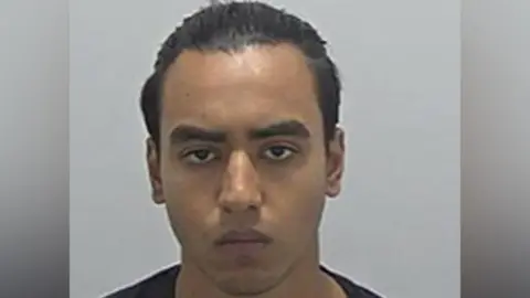 Bedfordshire Police Nazim Uddin's police custody photo, looking straight at the camera. He has short, slicked back, dark hair. He is wearing a dark top. 